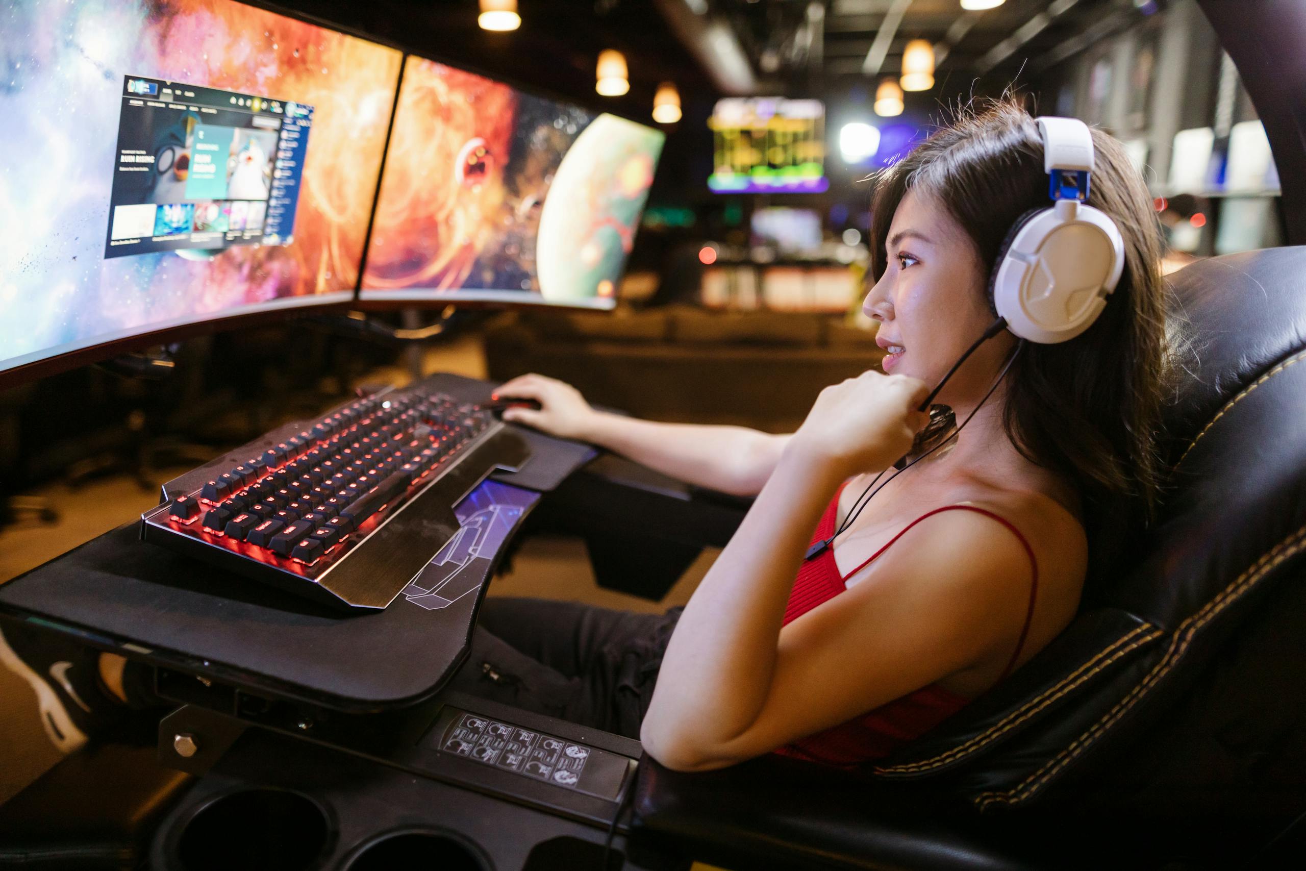 Young woman in red top gaming on dual monitors with white headphones in a modern setup.