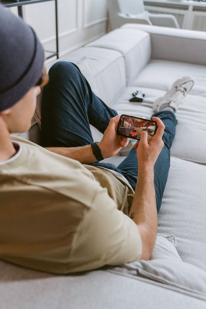 Young man wearing beanie playing a game on a smartphone while relaxing on a couch indoors.